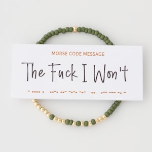 May include: A beaded bracelet with olive green and gold beads. A white card with the text "The Fuck I Won't" and Morse code is placed in the center of the bracelet. The bracelet is on a white background.