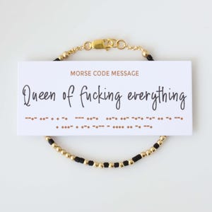 May include: A gold and black beaded bracelet with a card that reads "Queen of fucking everything" in cursive. The card also has the message in Morse code. The bracelet has a gold clasp.