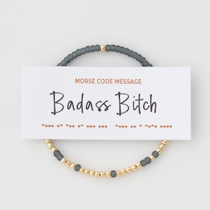 May include: A bracelet with grey and gold beads, presented with a card. The card displays the words "Badass Bitch" in a handwritten style, accompanied by Morse code symbols. The bracelet is a stylish accessory.