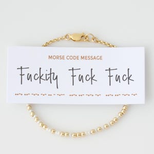 May include: Gold-toned beaded bracelet with a white card that reads "Fuckity Fuck Fuck" in black script. The card also says "Morse Code Message" and has a Morse code pattern below the text. The bracelet has a gold clasp.