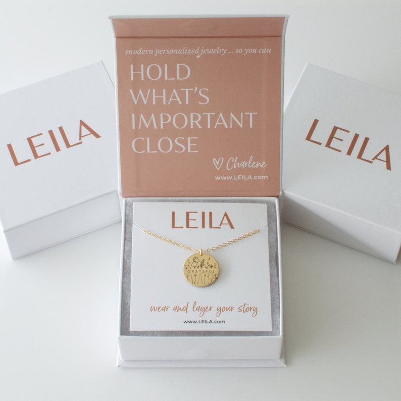 May include: A gold necklace with a round pendant that says "LEILA" and has a floral design. The necklace is in a white box with a pink lid that says "HOLD WHAT'S IMPORTANT CLOSE" and "Charlene" with the website address "www.LEILA.com".