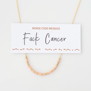 Peut inclure: Un collier doré avec une section perlée de couleur pêche. Le collier est associé à une carte blanche sur laquelle on peut lire "MORSE CODE MESSAGE" et "Fuck Cancer" en écriture noire. Des points et des tirets en code Morse se trouvent sous le texte.
