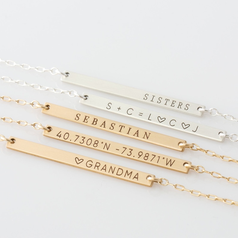 Puede incluir: Cuatro collares de barra de oro y plata con diferentes inscripciones. El collar superior dice "SISTERS S+C=L♡C♡J". El segundo collar dice "SEBASTIAN". El tercer collar dice "40.7308'N -73.9871'W". El collar inferior dice "♡GRANDMA".