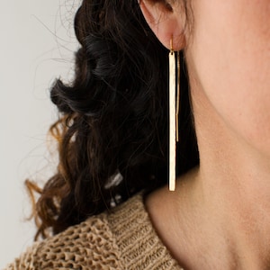 May include: Gold long bar earrings with a delicate chain detail hanging from the bottom.