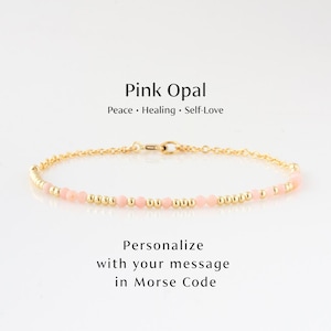 May include: A delicate gold bracelet featuring pink opal beads. The bracelet is accented with small gold beads and a clasp. Text on the image reads "Pink Opal, Peace, Healing, Self-Love" and "Personalize with your message in Morse Code."