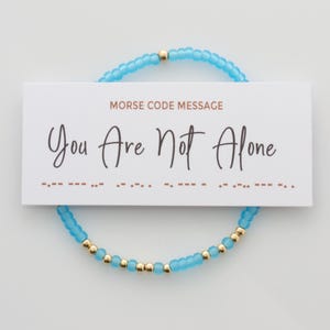May include: A blue and gold beaded bracelet with the message "You Are Not Alone" in Morse code on a white card.