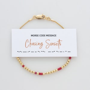 May include: A gold-colored bracelet with a series of small, round beads and a few square, red beads. A white card with the words "MORSE CODE MESSAGE" and "Chasing Sunsets" is attached to the bracelet.