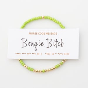 May include: A beaded bracelet with a lime green color and gold-colored beads. A white card with the text "Bougie Bitch" and "Morse Code Message" is placed in the center of the bracelet.