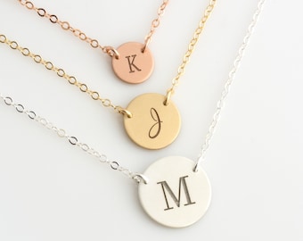 Puede incluir: Tres collares de oro con colgantes redondos que muestran las letras K, J y M. Los collares están superpuestos sobre un fondo blanco.