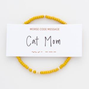 May include: A circular bracelet made of small yellow beads with a single gold-toned bead at the top. A white rectangular card rests over the bracelet, featuring the text MORSE CODE MESSAGE, Cat Mom, and a sequence of Morse code dots and dashes.