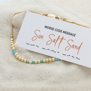 May include: A gold chain bracelet with turquoise beads and a card that says "Morse Code Message" with the words "Sun Salt Sand" written in cursive and the Morse code for each word below.