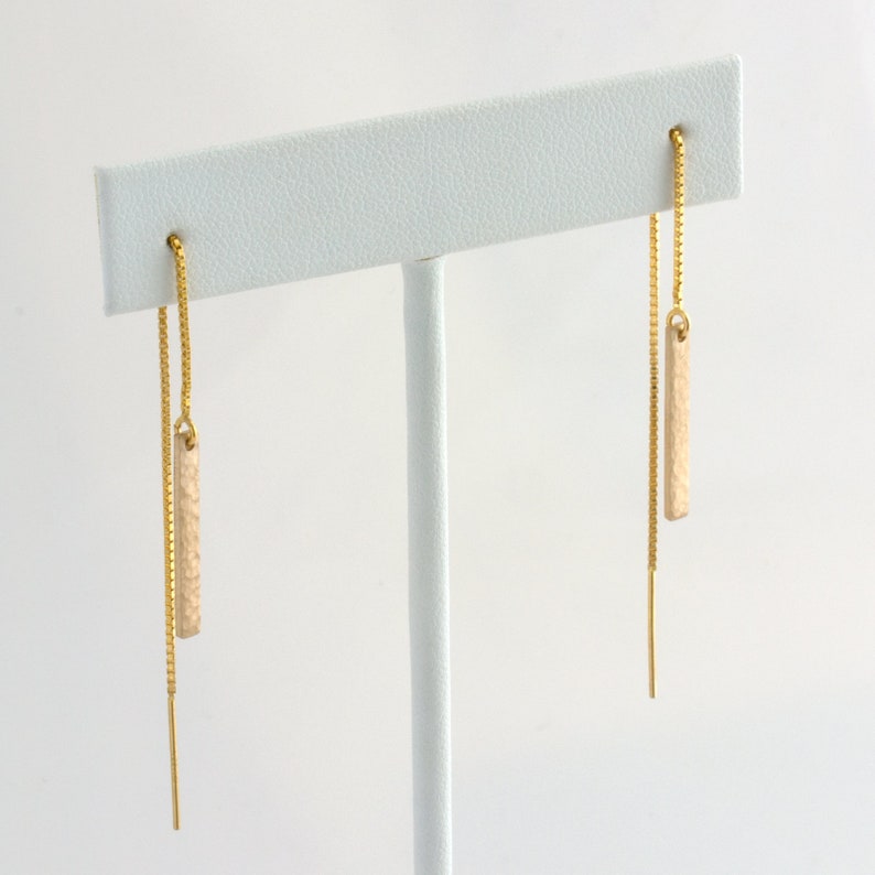Puede incluir: Un par de pendientes colgantes dorados. Cada pendiente presenta una delicada cadena dorada con un dije texturizado, rectangular y blanquecino. Los pendientes se exhiben en un soporte blanco para pendientes.