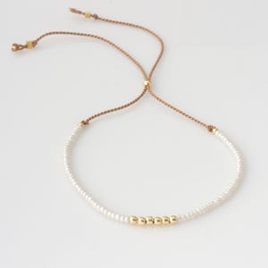 May include: Delicate white beaded bracelet with a sliding knot closure. The bracelet features a row of gold-colored beads and a tan cord. A simple and elegant piece of jewelry.