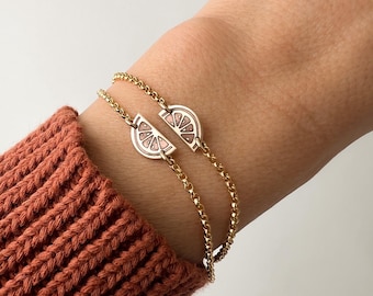 May include: Two gold chain bracelets with a pink and white enamel half-circle lemon slice charm on each. The charms are connected together.
