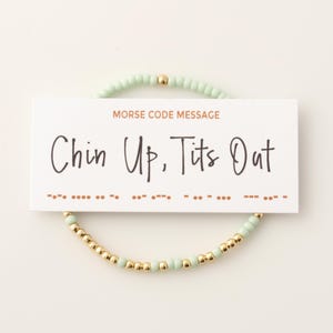 May include: A beaded bracelet with a card that reads "Chin Up, Tits Out." The bracelet features light green and gold beads. The card also says "Morse Code Message."