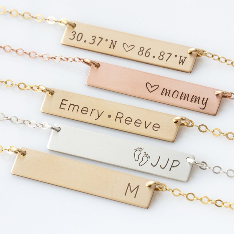 Op de afbeelding: Vijf gouden bar kettingen met verschillende gravures. De bovenste ketting heeft de co&ouml;rdinaten "30.37&deg;N &hearts; 86.87&deg;W". De tweede ketting zegt "mama". De derde ketting zegt "Emery &bull; Reeve". De vierde ketting heeft een voetafdruk symbool en de initialen "JJP". De vijfde ketting heeft de letter "M" gegraveerd.