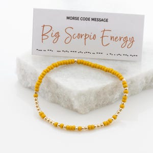 May include: A yellow beaded bracelet with gold beads and a card that reads "Morse Code Message Big Scorpio Energy".