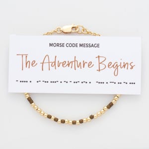May include: A gold bracelet with brown beads that spells out "The Adventure Begins" in Morse code. The bracelet is on a white card with the message "Morse Code Message" and the phrase "The Adventure Begins" written in brown.