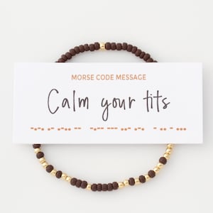 May include: A beaded bracelet with brown and gold beads encircles a white card. The card reads "MORSE CODE MESSAGE" and "Calm your tits" in black script. Morse code dots and dashes are below the text.