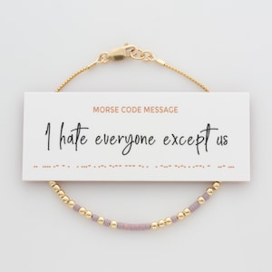 May include: A gold-coloured beaded bracelet with a message card. The card reads "I hate everyone except us" in cursive, with a Morse code representation below. The bracelet has gold beads and lavender-coloured beads.