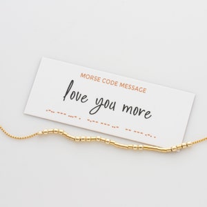 May include: A white card with the text "Morse Code Message" and the phrase "love you more" written in black cursive. Below the phrase is a line of dots and dashes representing the message in Morse code.
