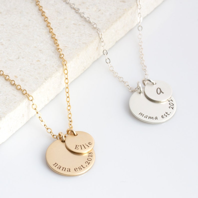 May include: Two gold and silver necklaces with personalised discs. The gold necklace has two discs, one with the name "Ellie" and the other with "nana est. 2018". The silver necklace has two discs, one with the letter "A" and the other with "mama est. 2017".
