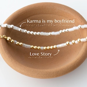 May include: Two beaded necklaces with the phrases "Karma is my boyfriend" and "Love Story". The top necklace has silver beads and the bottom necklace has gold beads. The necklaces are displayed in a small, brown, terracotta bowl.