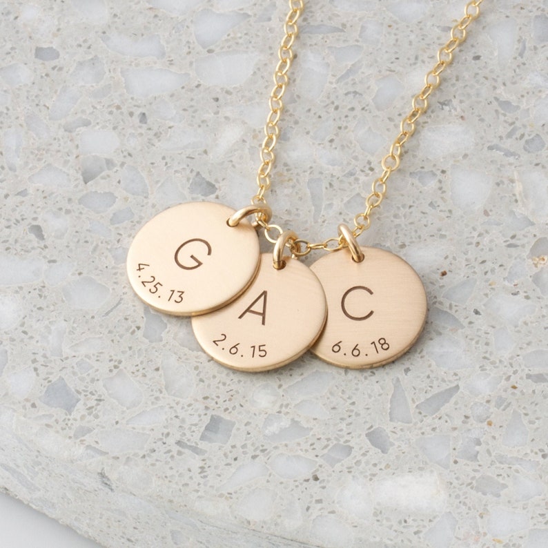 May include: Gold necklace with three round pendants. Each pendant has a letter engraved on it, G, A, and C, with dates underneath.  The dates are 4.25.13, 2.6.15, and 6.6.18.