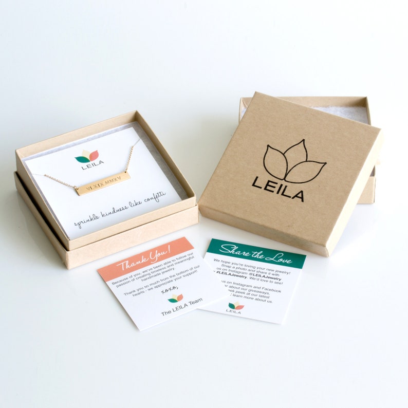 May include: A gold necklace with a bar pendant that says "VLXIX MMXV" in a simple font. The necklace is in a small brown box with a green logo that says "LEILA". The box is on a white surface with two cards that say "Thank You!" and "Share the Love".