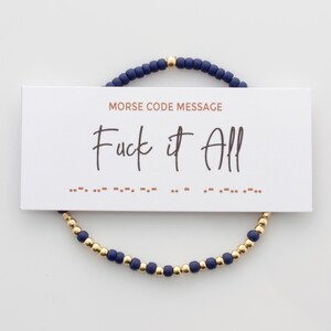 May include: A navy blue and gold beaded bracelet with a card that reads "Morse Code Message" and "Fuck it All" in cursive. The card also includes the Morse code for the message.