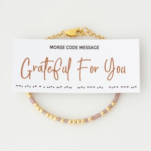 May include: A gold-colored beaded bracelet with a card that reads "MORSE CODE MESSAGE Grateful For You". The bracelet features small gold and lavender beads. The bracelet is connected with a gold chain and clasp.
