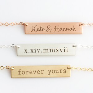 Op de afbeelding: Drie gouden bar kettingen met verschillende gegraveerde berichten. De bovenste ketting heeft het bericht "Kate & Hannah" gegraveerd. De middelste ketting heeft het bericht "x.xiv.mmxvii" gegraveerd. De onderste ketting heeft het bericht "forever yours" gegraveerd.