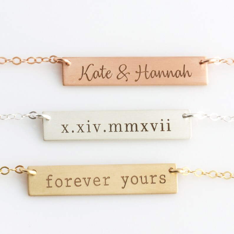 Op de afbeelding: Drie gouden bar kettingen met verschillende gegraveerde berichten. De bovenste ketting heeft het bericht "Kate & Hannah" gegraveerd. De middelste ketting heeft het bericht "x.xiv.mmxvii" gegraveerd. De onderste ketting heeft het bericht "forever yours" gegraveerd.