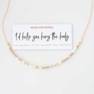 May include: A gold-coloured necklace with a Morse code design. The necklace has small white and gold beads. A white card with the text "I'd help you bury the body" is behind the necklace.