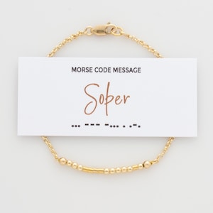 May include: Gold chain bracelet with small beads and a bar charm. The bracelet has a message in Morse code that reads "Sober".