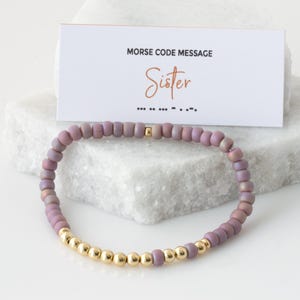 May include: A purple and gold beaded bracelet with a card that reads "Morse Code Message Sister" and the Morse code for the word "Sister".