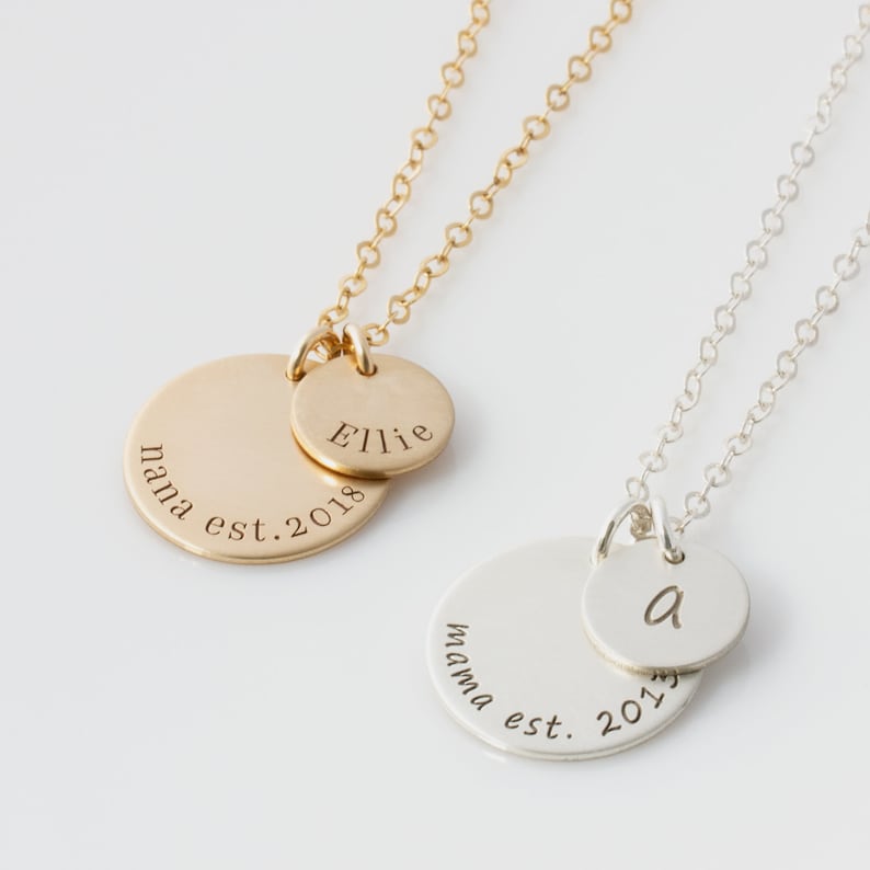 May include: Two gold and silver necklaces with round pendants. The gold pendant has the text "nana est. 2018" and a smaller pendant with the text "Ellie". The silver pendant has the text "mama est. 2017" and a smaller pendant with the letter "a".