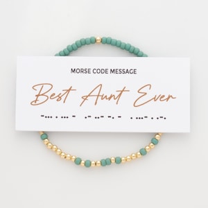 May include: A beaded bracelet with a card that reads "Best Aunt Ever." The bracelet has two strands, one with gold beads and teal beads, and the other with teal beads. The card says "Morse Code Message."
