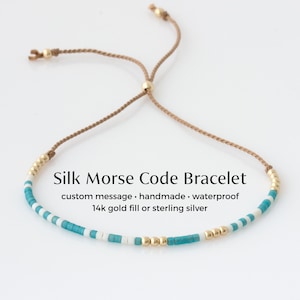 May include: A silk Morse code bracelet with turquoise, white, and gold beads. The adjustable bracelet has a sliding knot closure and the text "Silk Morse Code Bracelet". The bracelet is handmade and waterproof.
