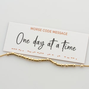 May include: A gold bracelet with a card that reads "Morse Code Message" and "One day at a time". The bracelet is made of small gold beads.