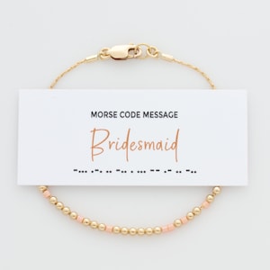 May include: Gold-colored beaded bracelet with a lobster clasp, featuring a Morse code message. A white card with the word "Bridesmaid" in a copper-colored script is attached to the bracelet. The bracelet has gold and peach-colored beads.