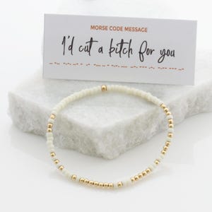 May include: A white beaded bracelet with gold-colored accents. A card with the text "MORSE CODE MESSAGE" and "I'd cut a bitch for you" is behind the bracelet. The bracelet is on a white surface.