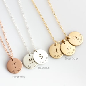 May include: Three gold necklaces with round pendants. Each pendant has a letter and a date engraved on it. The necklaces are on a white background. The engraving styles are handwriting, typewriter, and brush script.
