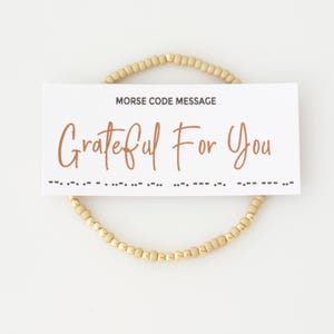 May include: A circular beaded bracelet featuring alternating matte tan and metallic gold beads. A white rectangular card is placed over the bracelet, displaying the text MORSE CODE MESSAGE and Grateful For You in brown, with Morse code dots and dashes.