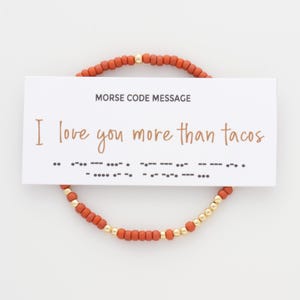 May include: A circular beaded bracelet featuring matte burnt orange beads and small metallic gold beads. A white rectangular card rests on top, displaying the text MORSE CODE MESSAGE, I love you more than tacos, and the corresponding Morse code dots and dashes.