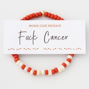 May include: A bracelet made of red and white beads, with a gold-coloured bead. A white card with the text "MORSE CODE MESSAGE" and "Fuck Cancer" is in front of the bracelet. The bracelet is set against a white backdrop.