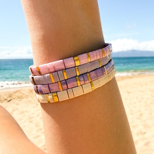 May include: Four colorful tile bracelets in shades of pink, lavender, and cream with gold accents are stacked on a wrist. The bracelets are rectangular and appear to be made of ceramic or a similar material. The background shows a beach and ocean.