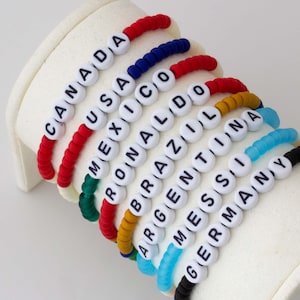 Peut inclure: Un empilement de huit bracelets de perles composés de disques en argile colorés et de perles à lettres blanches formant les mots CANADA, USA, MEXICO, RONALDO, BRAZIL, ARGENTINA, MESSI et GERMANY, disposés sur un présentoir blanc incurvé.