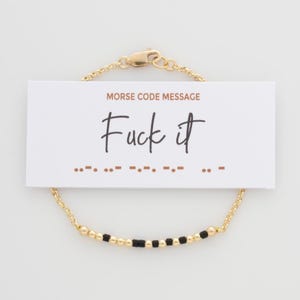 May include: A gold-colored bracelet with a Morse code design. The bracelet is paired with a white card that reads "Morse Code Message" and "Fuck it" in black script. The Morse code is represented by black and gold beads.