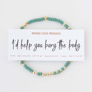 May include: A teal beaded bracelet with gold accents encircles a white card. The card reads "I'd help you bury the body" in cursive, with a Morse code message below. The bracelet is a unique, quirky accessory.